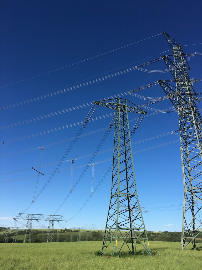 Hochspannungsleitungen auf einem grünen Feld unter blauem Himmel, im Vordergrund rechts zwei große Masten und Leitungen, im Hintergrund weitere Masten und Leitungen