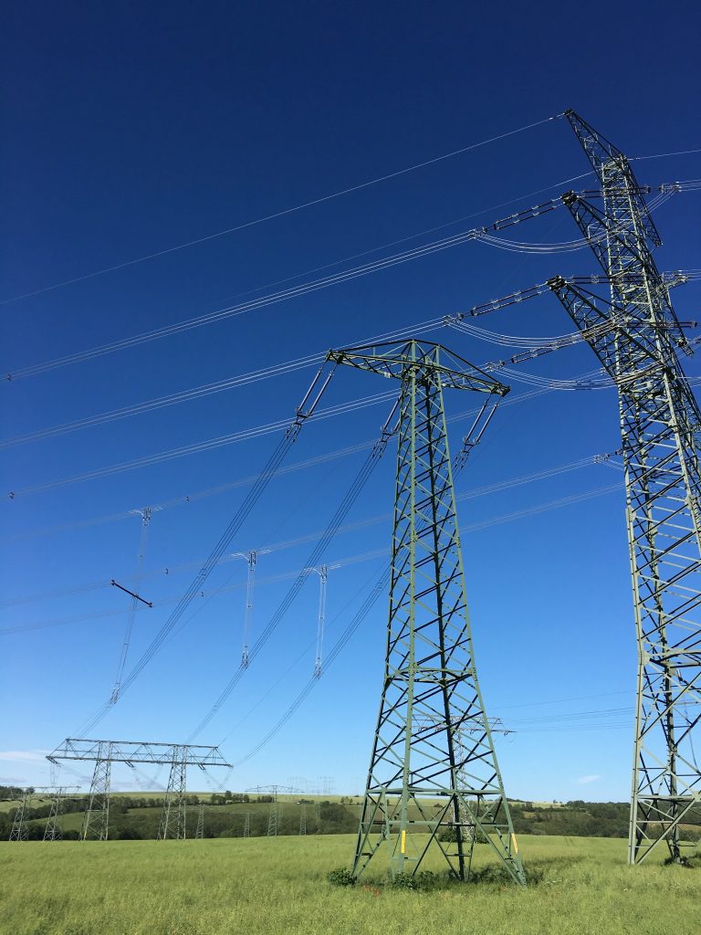 Power lines on a green field under a blue sky, in the foreground on the right two large pylons and lines, in the background more pylons and lines