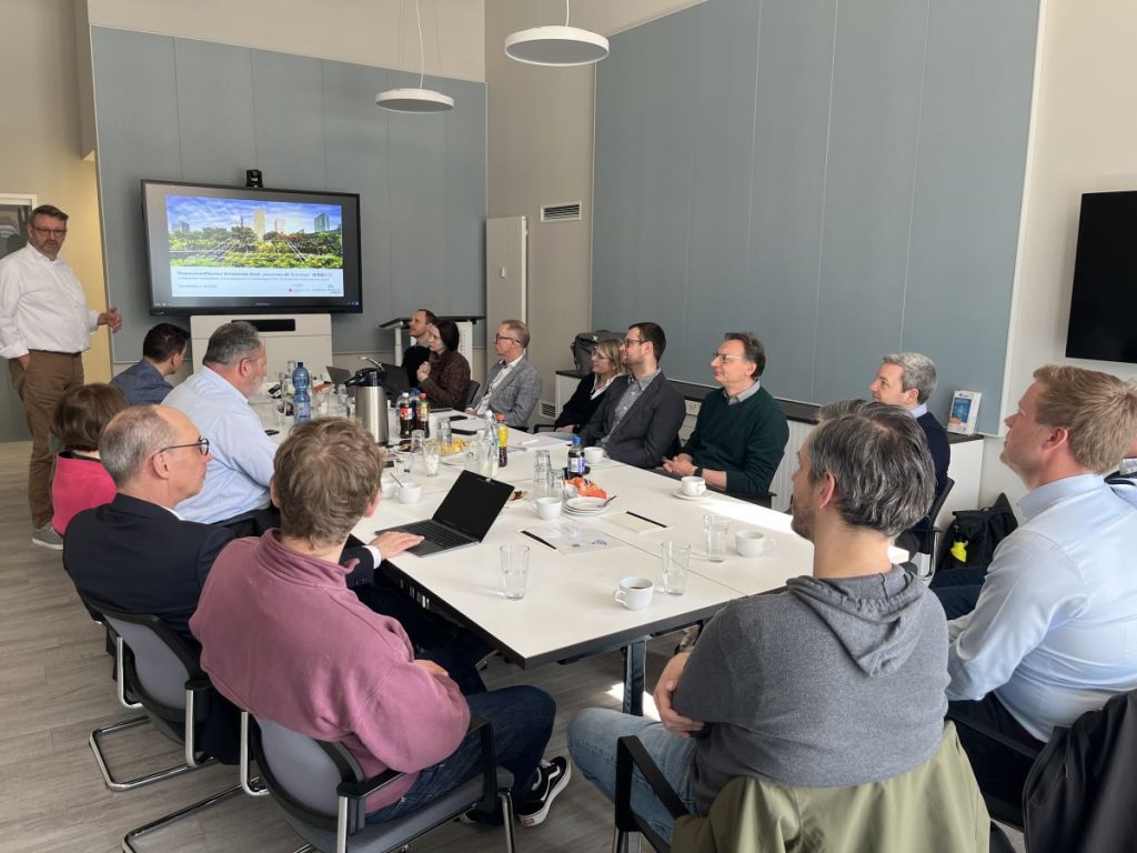 15 Personen sitzend an einem Konferenztisch, links hinten eine Person stehend neben einem Bildschirm mit Präsentation