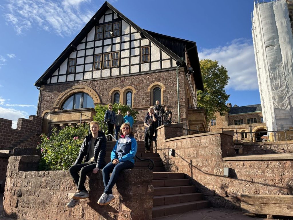 Neun Personen an und auf einer hellbraunen Treppe vor einem Gebäude der Wartburg mit Fachwerk-Dach
