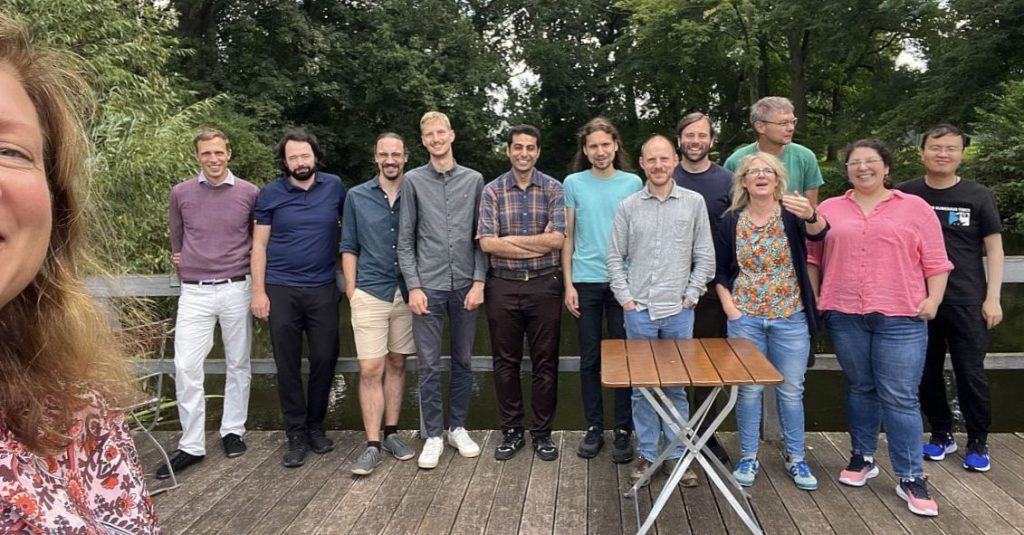 Teamfoto mit zwölf Personen und einer Person links in Selfie-Perspektive, im Vordergrund Holzplanken, im Hintergrund Gewässer und Grün