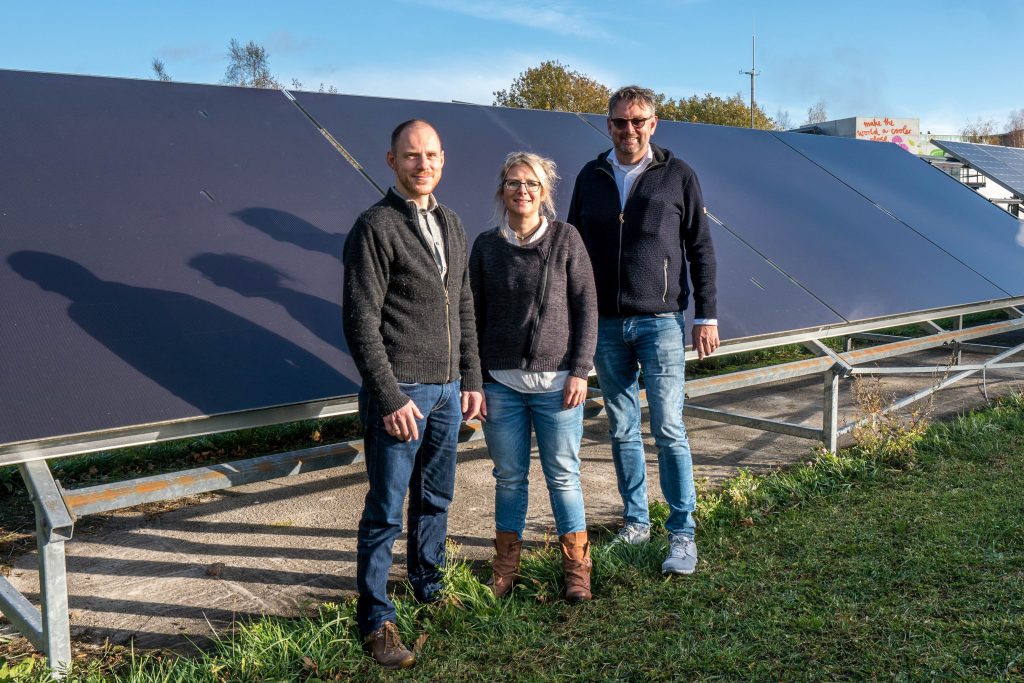 two male and one female scientist (in the middle) standing in front of elevated photovoltaic modules, in the foreground lawn, in the background trees and buildings