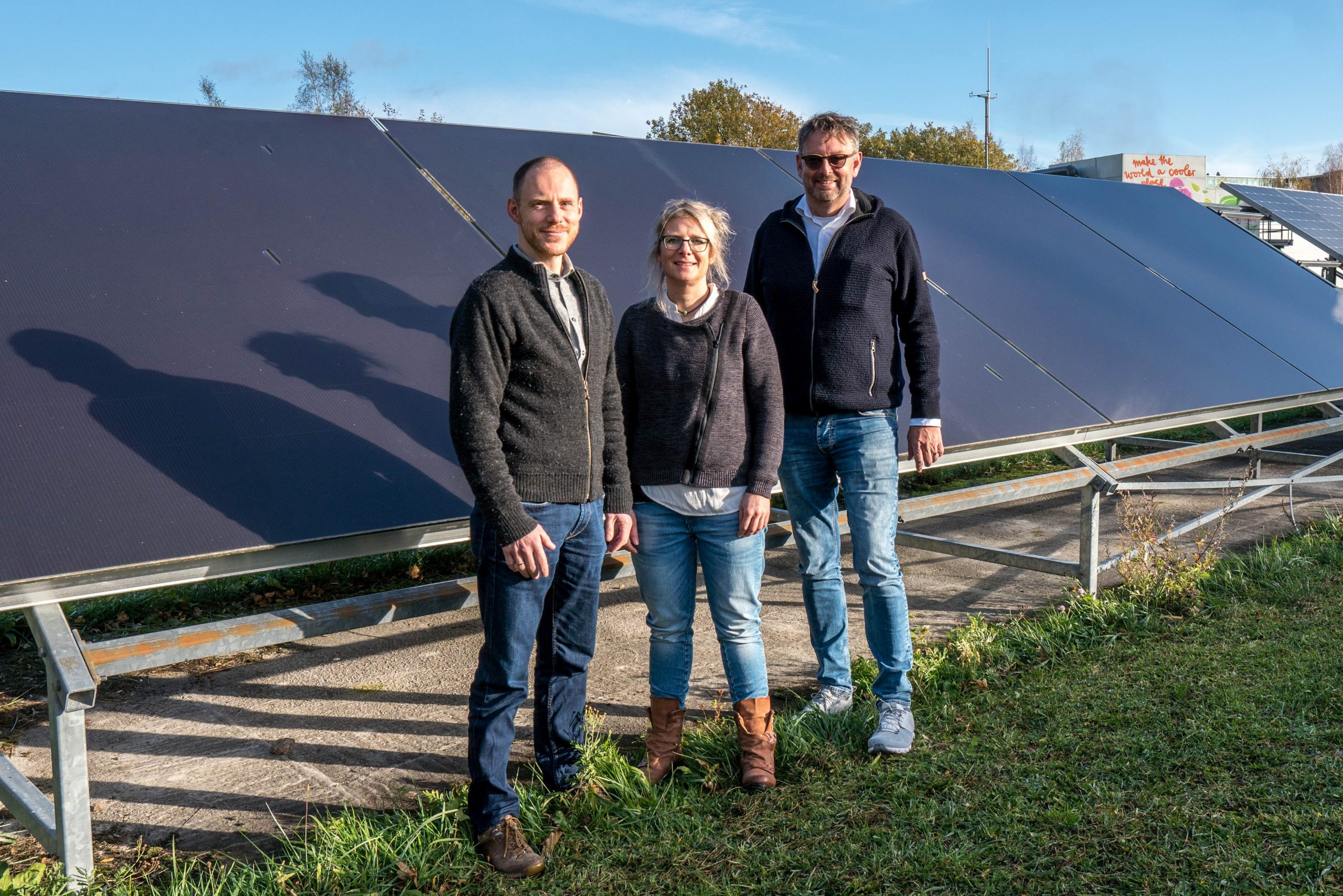 zwei männliche und eine weibliche Wissenschaftlerin (in der MItte) stehend vor aufgeständerten Photovoltaikmodulen, im Vordergrund Rasen, im Hintergrund Bäume und Gebäude